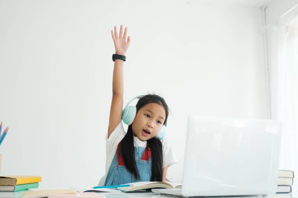 Young student raising hand during an online class, using a creative suite on her laptop for an interactive learning experience.