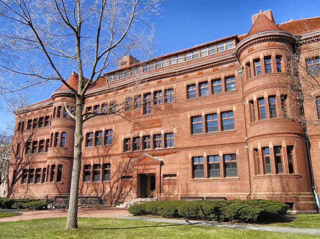 Historic red brick school building with a lush green lawn and a bare tree in the foreground under a clear blue sky.